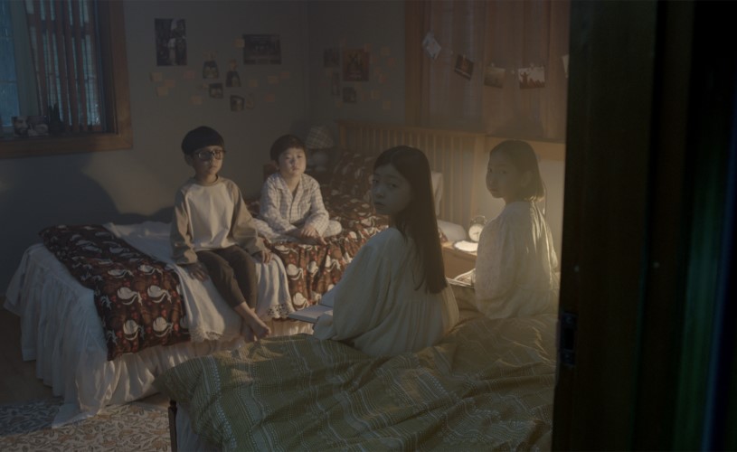 Two children sat on a bed and another standing, all looking to camera